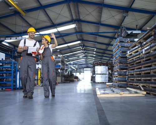 Factory workers in work wear and yellow helmets walking through industrial production hall and discussing about organization.